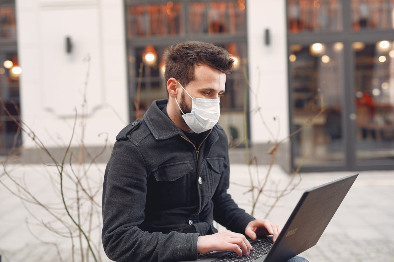 Young bearded male entrepreneur wearing warm jacket and protective facial mask during remote work against city building exterior in cold season